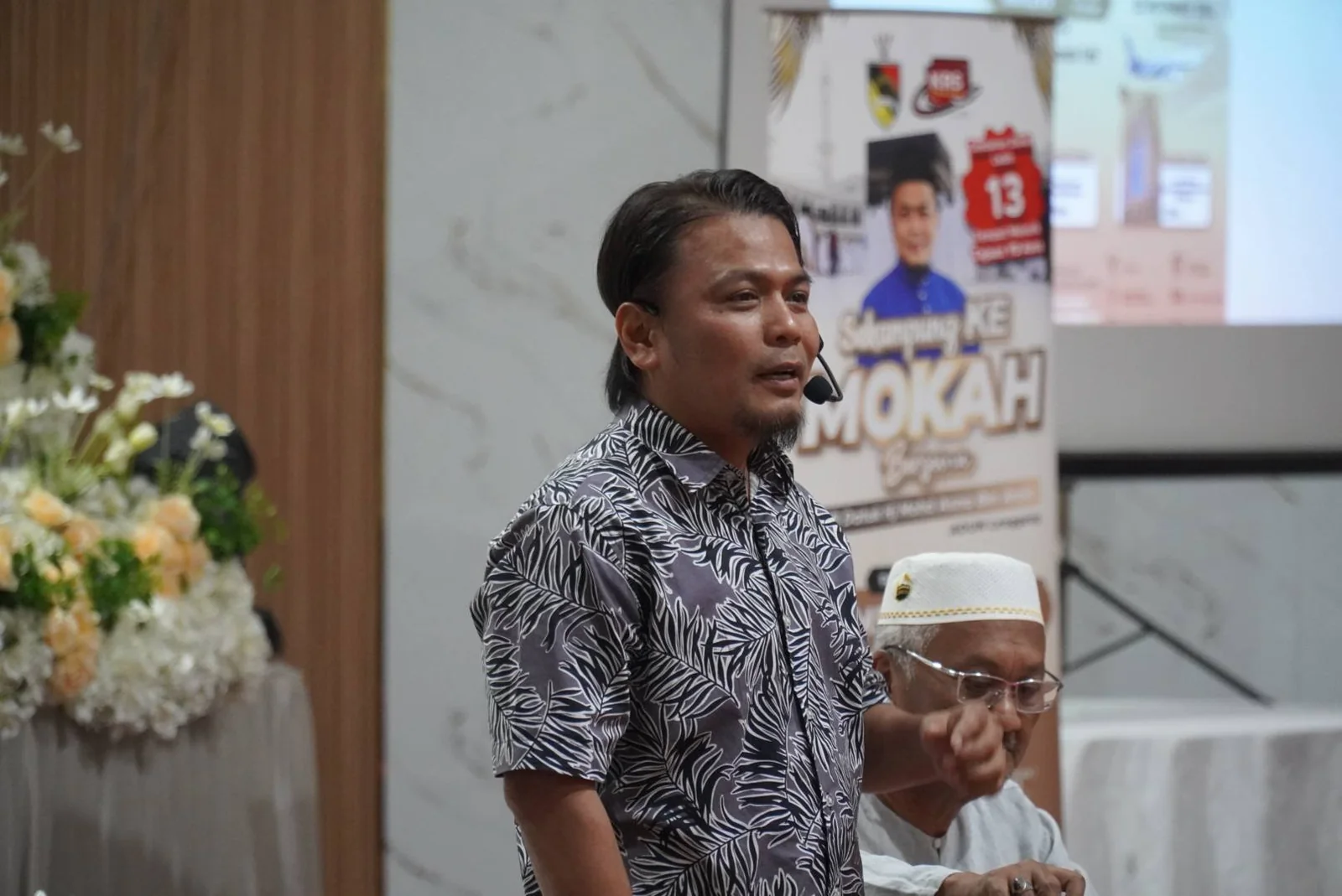 Penceramah menyampaikan taklimat kursus umrah dengan poster Sekampung Ke Mokah dipaparkan di skrin