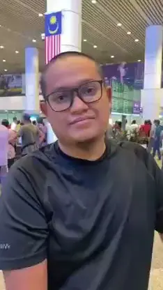 Jemaah lelaki muda bercermin mata memberi testimoni di balai berlepas berbendera Malaysia