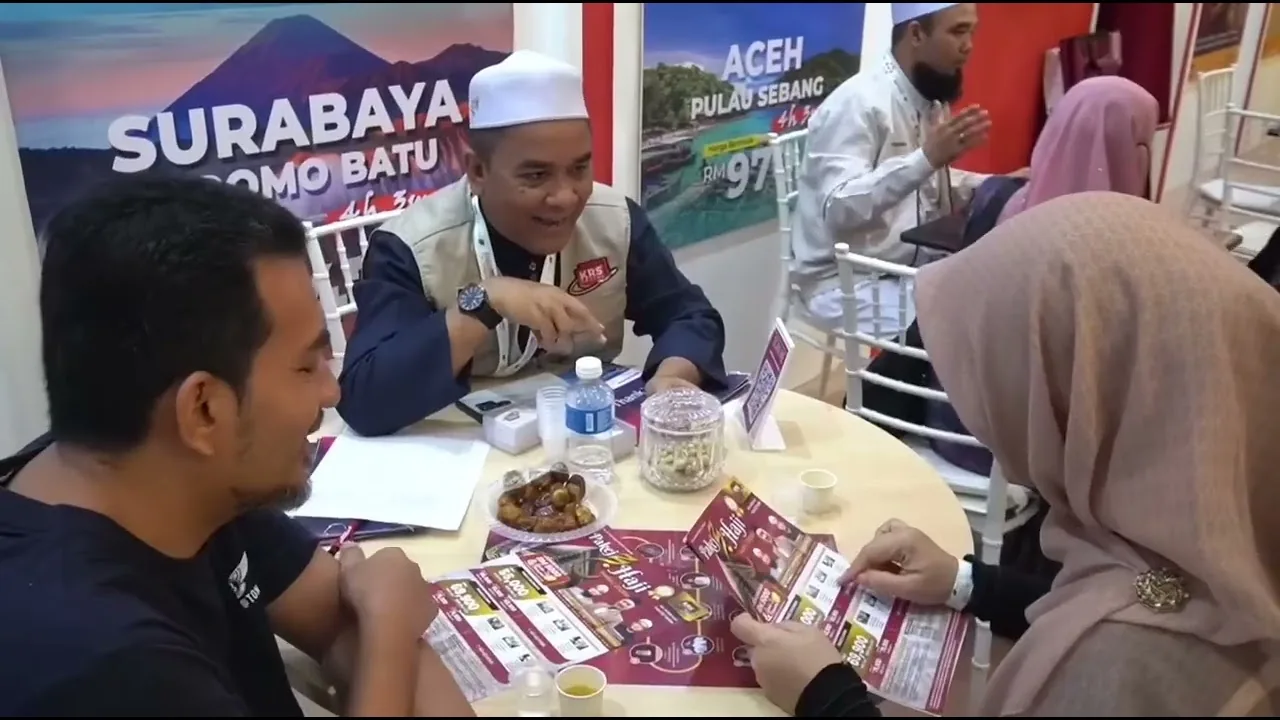 Petugas KRS Travel berbual dengan bakal jemaah di meja pameran, dengan poster pakej pelancongan Surabaya dan Aceh di belakang