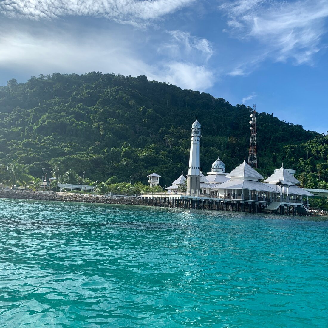 Masjid putih berkubah di atas tiang menghadap air pirus Pulau Perhentian, dengan bukit berhutan di latar, Terengganu — Pakej Pulau Perhentian KRS Travel