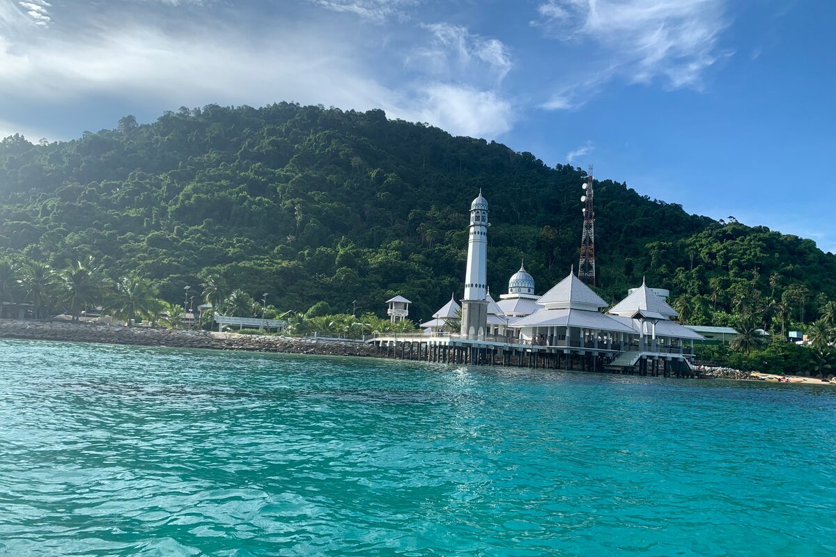 Masjid putih berkubah di atas tiang menghadap air pirus Pulau Perhentian, dengan bukit berhutan di latar, Terengganu — Pakej Pulau Perhentian KRS Travel